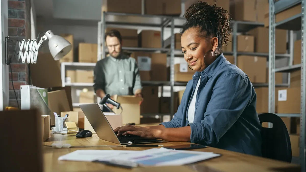 Woman working on computer with man in back carrying a box